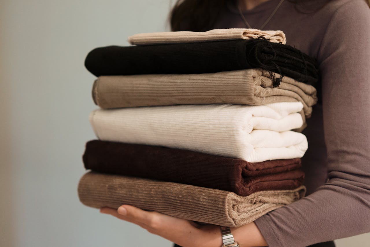 A person holds neatly folded corduroy fabrics in various neutral colors, offering a cozy and warm aesthetic.